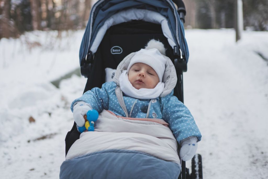 En bebis klädd i vinterkläder ligger bekvämt i en åkpåse i en barnvagn på en snöig stig.