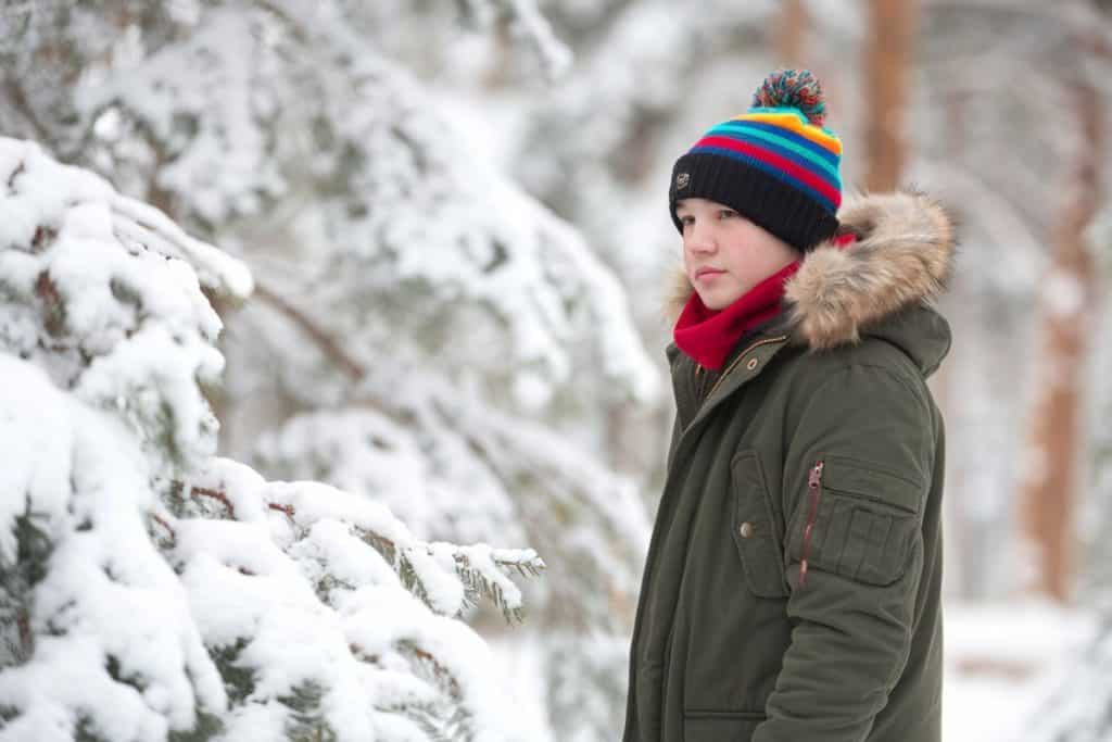 Ett barn klädd i en varm grön parkas med pälsfodrad huva och en färgglad mössa står i en snöig skog.
